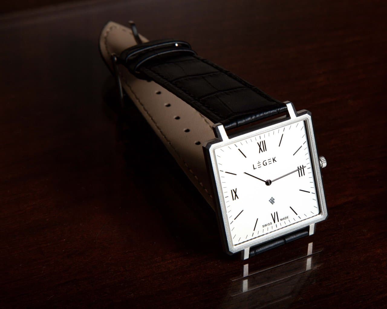 Product photography of a square silver Swiss watch with a black leather strap on dark wood