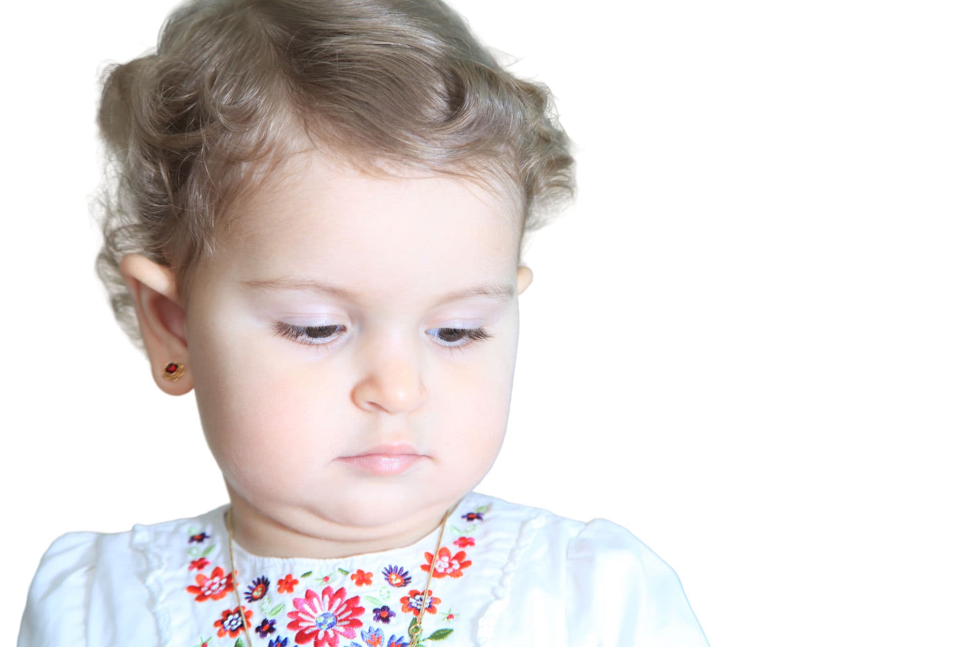 Portrait of a baby girl with curly hair in an embroidered white top, looking down, on a white background